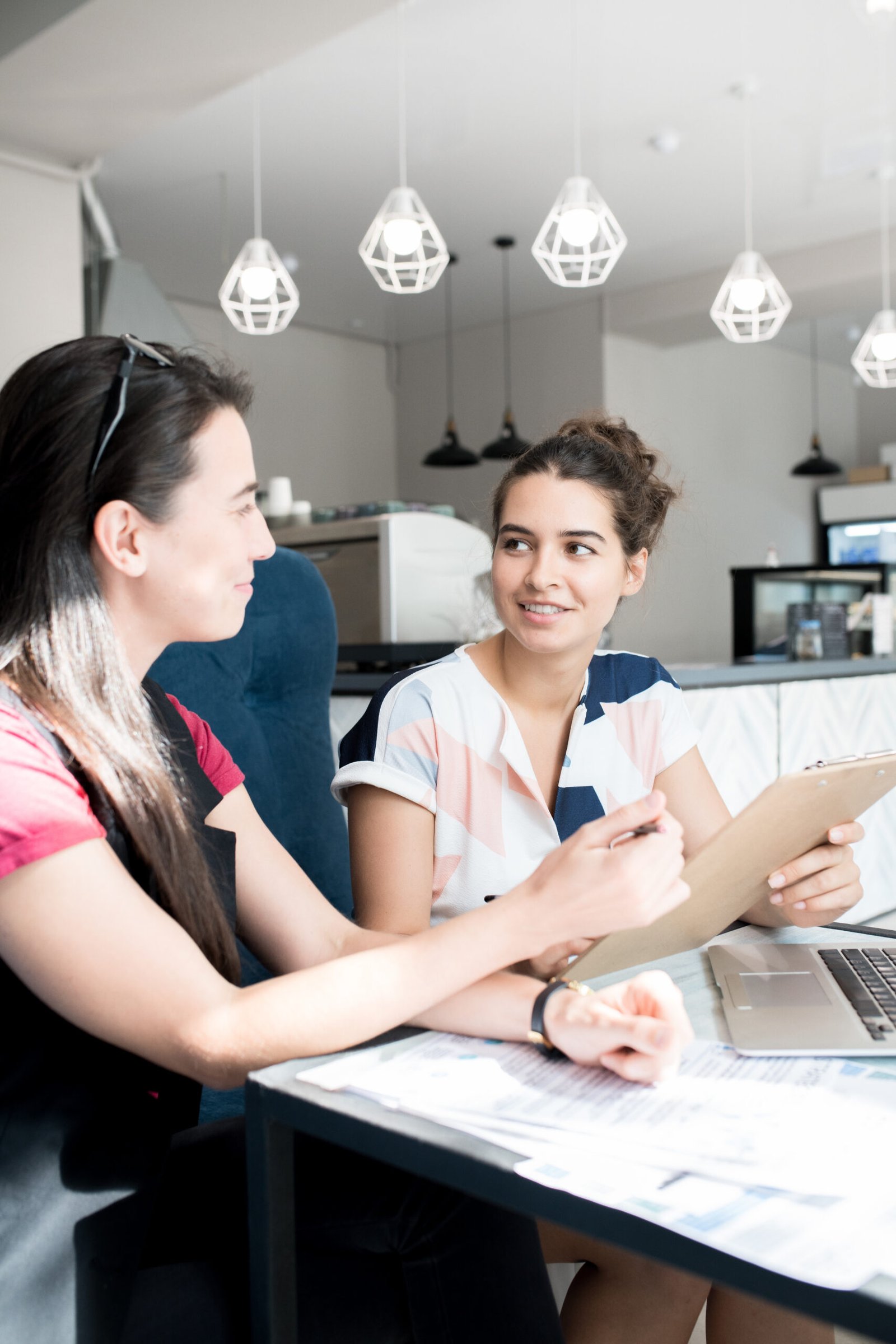 Two women engaged in a discussion at a cafe table, reviewing documents and a laptop, representing collaboration and communication in property appraisal services.